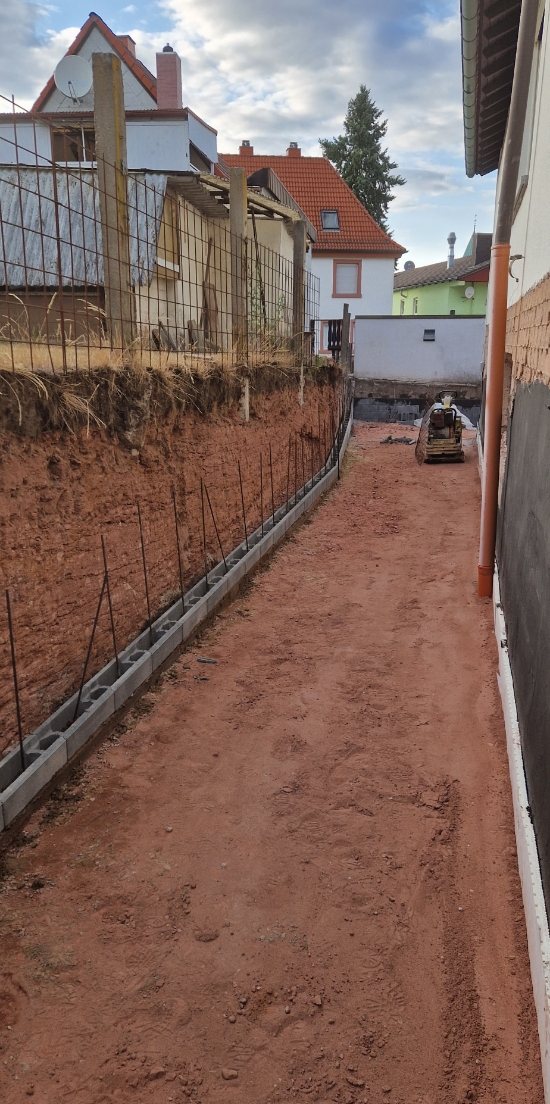 Schmaler, unbefestigter Weg zwischen Häuserwänden mit Bauarbeiten auf der linken Seite und einer kleinen Maschine am Ende.