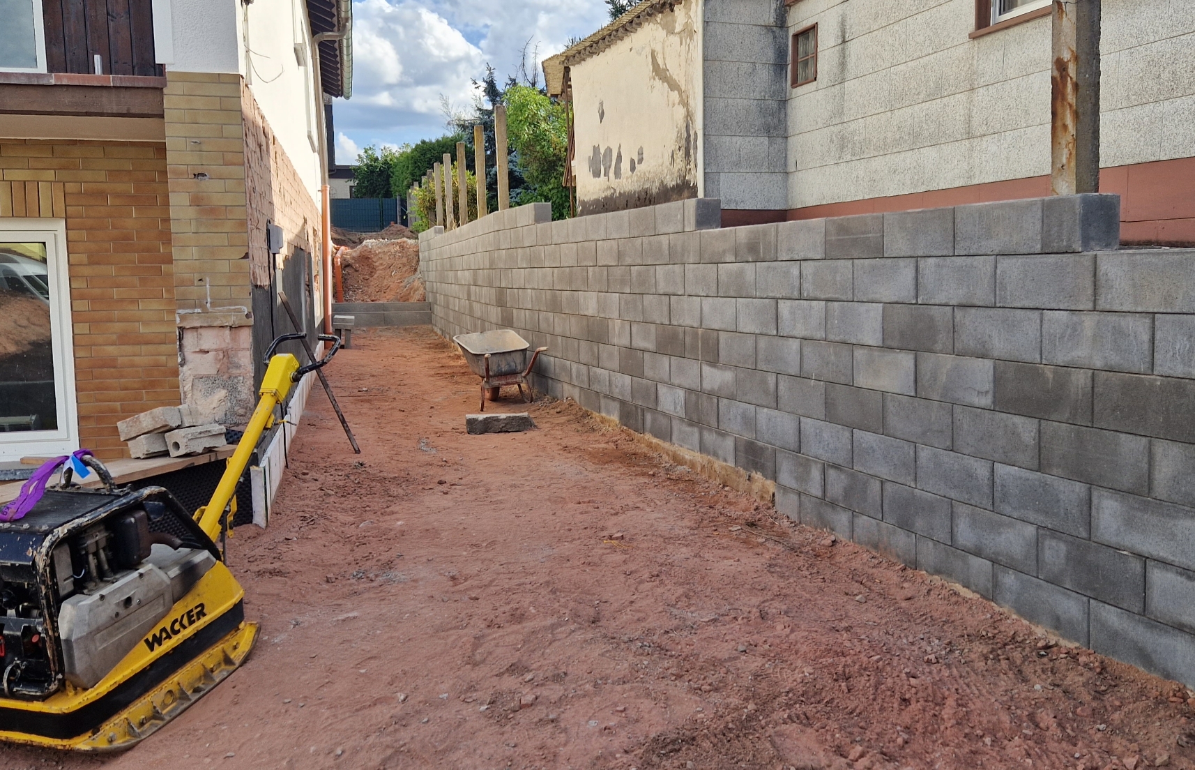 Baustelle zwischen zwei Häusern mit einer frisch errichteten grauen Betonmauer, einer Schubkarre und einer gelben Rüttelplatte auf unbefestigtem Boden.