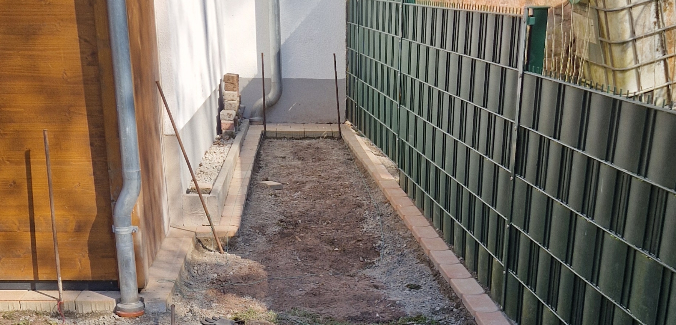 Schmaler Gartenweg mit frisch angelegtem Boden, umgeben von Backsteinkanten, einer Hauswand mit Fallrohr links und einem grünen Metallzaun rechts.