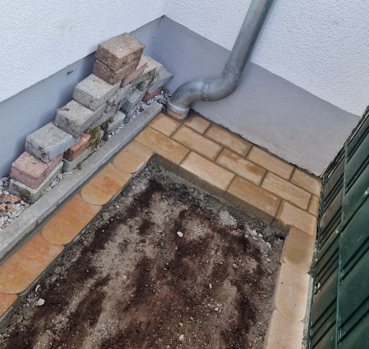 Bodenfläche mit Erde neben einer Hauswand, neu verlegt mit rechteckigen und runden Pflastersteinen, daneben stapelweise Ziegelsteine und ein Regenrohr.