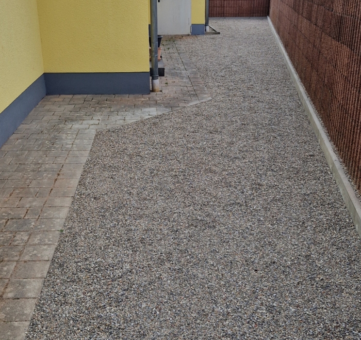 Gepflasterter und kiesbedeckter Weg neben einem gelben Hauswand mit einer braunen Sichtschutzwand rechts.