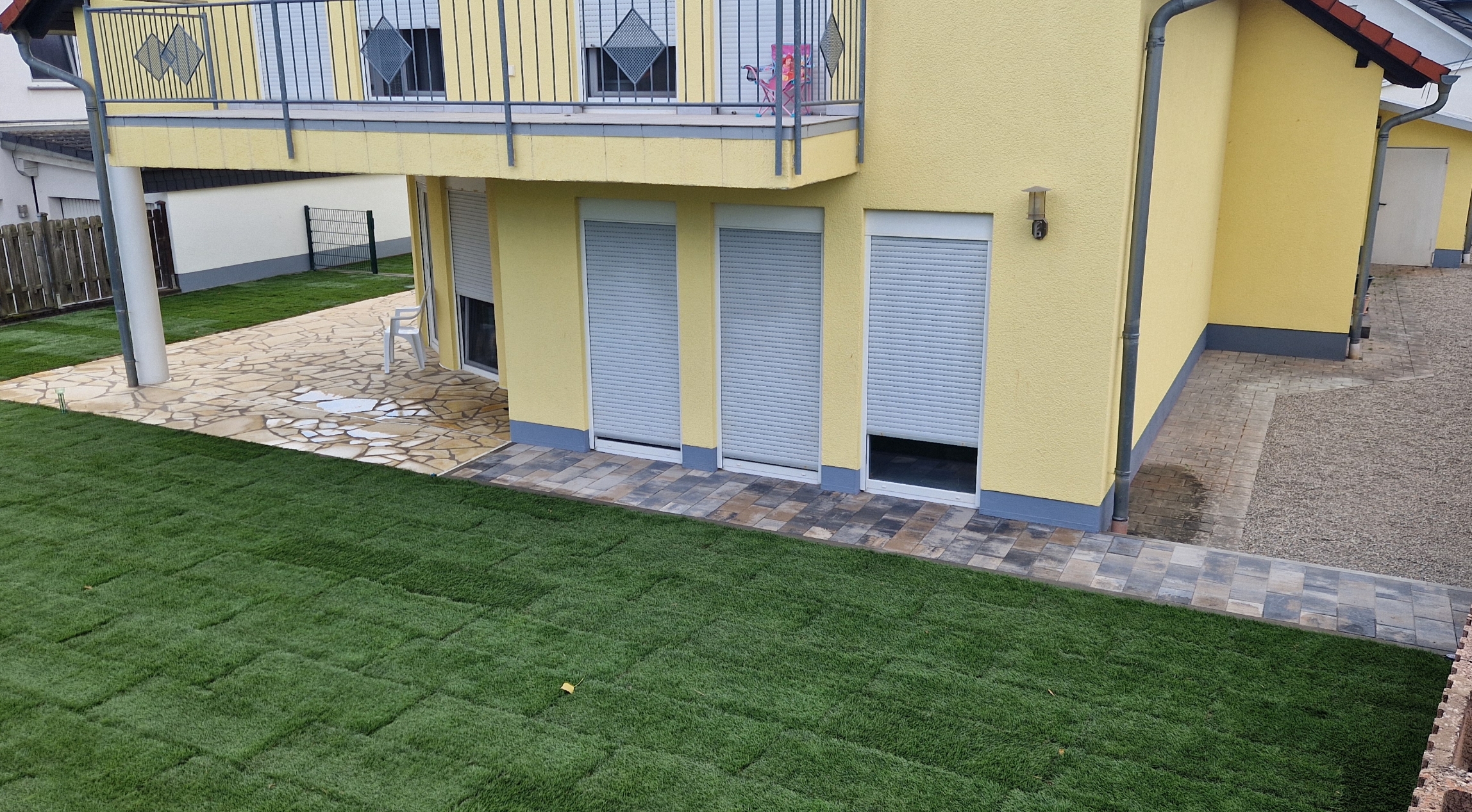 Garten mit frisch verlegtem grünem Rasen vor einem gelben Haus mit geschlossenen Fensterrollläden und einem Balkon.