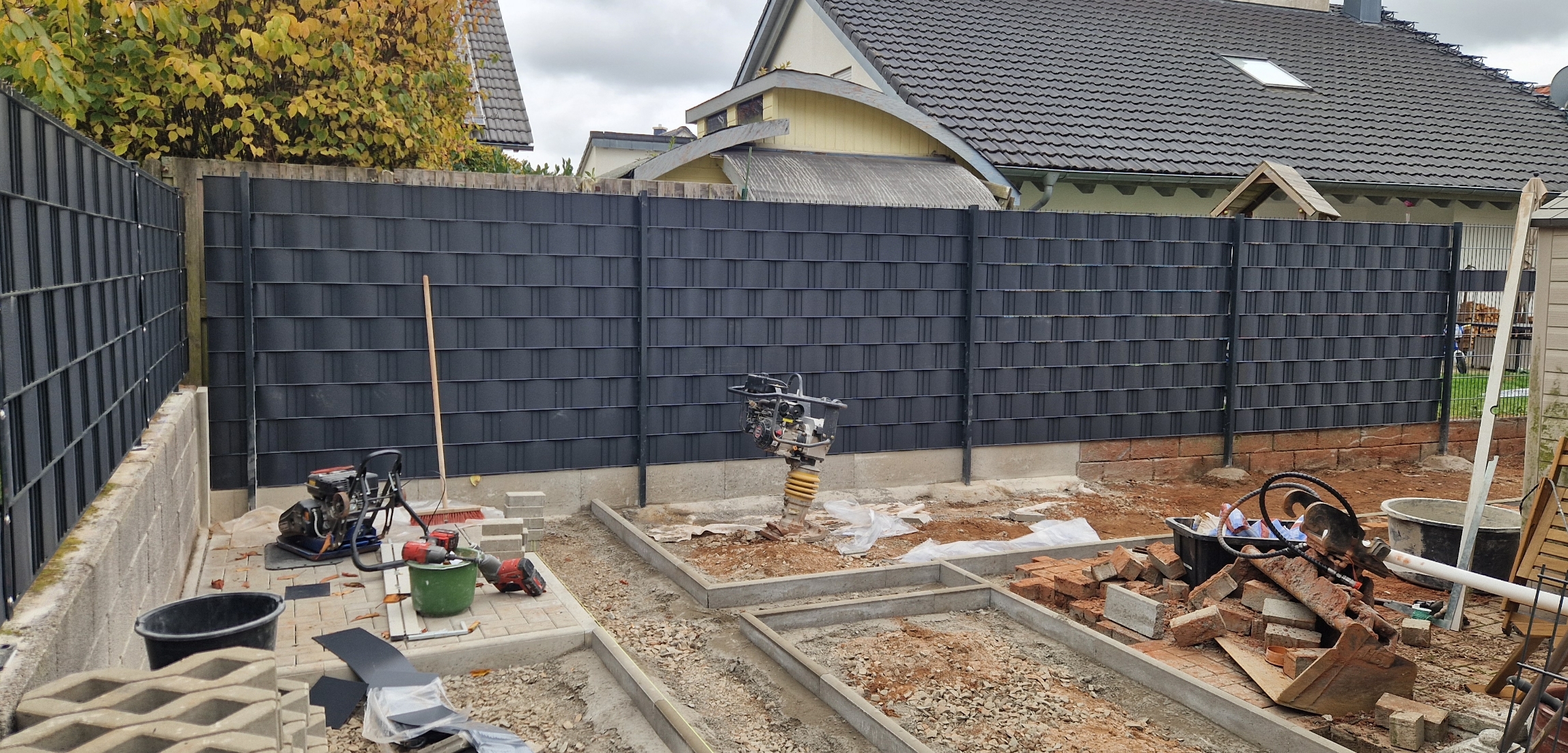 Baustelle im Garten mit Baustoffen, Baugeräten und schwarzem Zaun vor einem Haus.