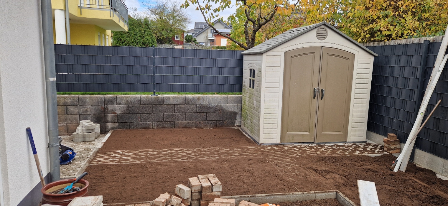 Kleiner Gartenbereich mit frisch geebnetem Erdreich, einem Hühnerstall rechts, umgeben von einer schwarzen Zaun- und Steinmauer.