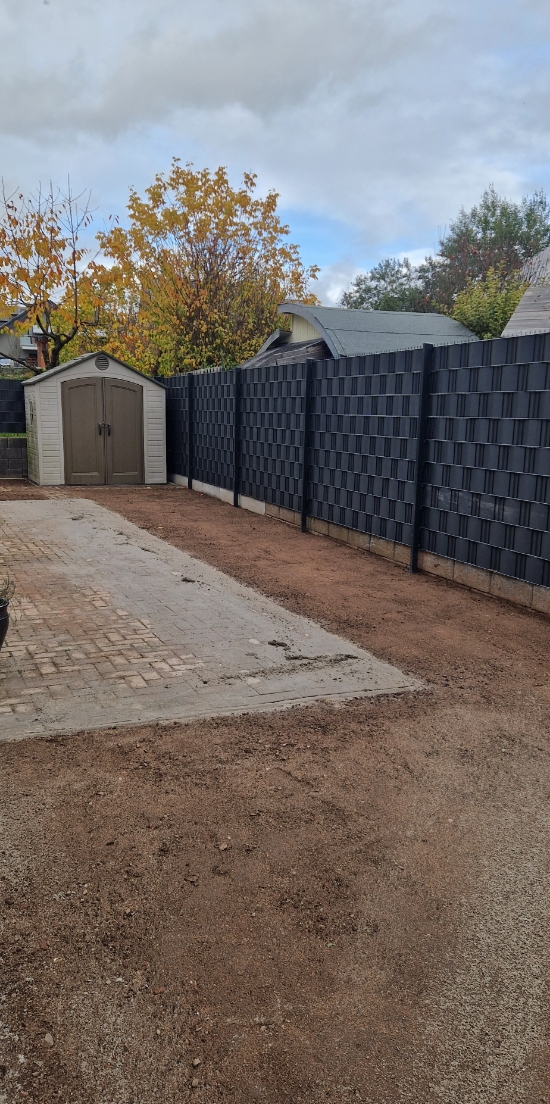 Ein Garten mit einem schmalen gepflasterten Weg, einem kleinen Geräteschuppen und einem hohen schwarzen Sichtschutzzaun, umgeben von herbstlichen Bäumen.