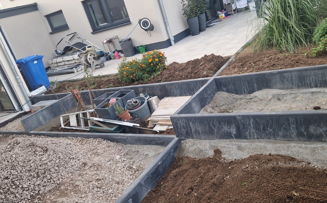 Hausgarten mit mehreren rechteckigen Beeten, einige gefüllt mit Erde und Kies, andere mit Blumenkübeln und Gartenwerkzeugen.