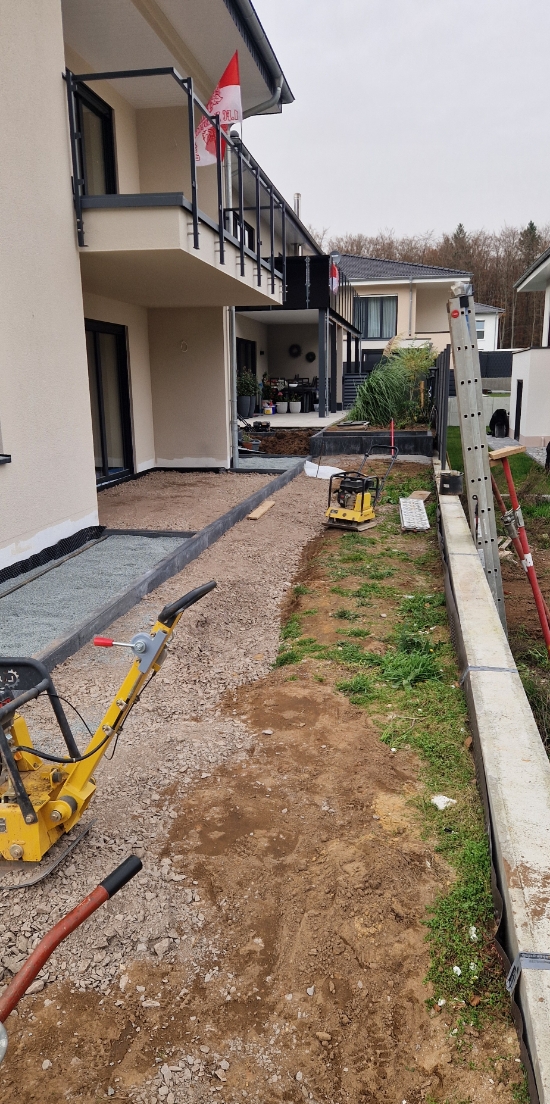 Baustelle mit gelben Rüttelplatten und Schubkarre neben modernem Wohngebäude mit Balkon und Garten in Bauphase.
