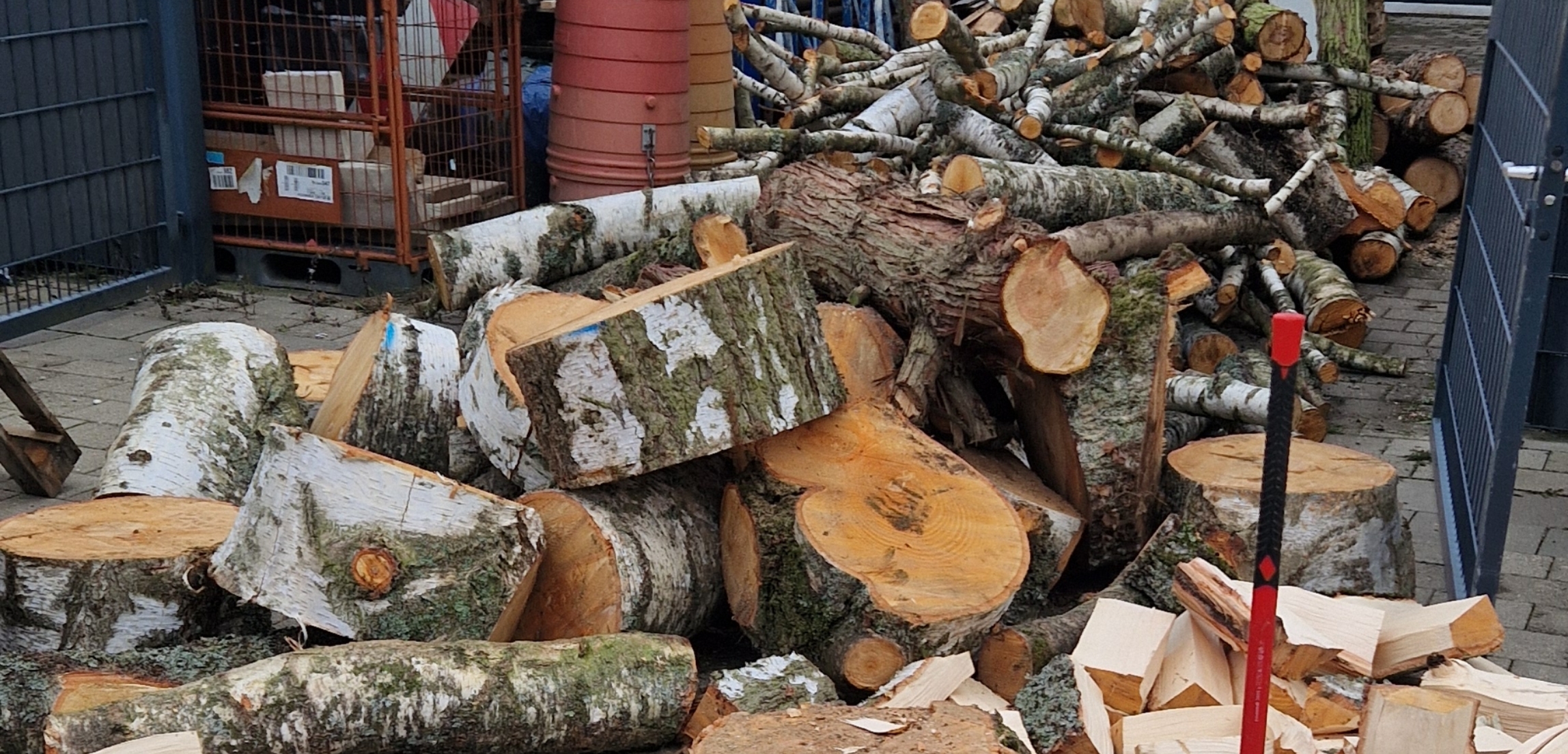 Großer Stapel geschnittener Birkenstämme und Äste, daneben ein Holzhacke mit rotem Griff.