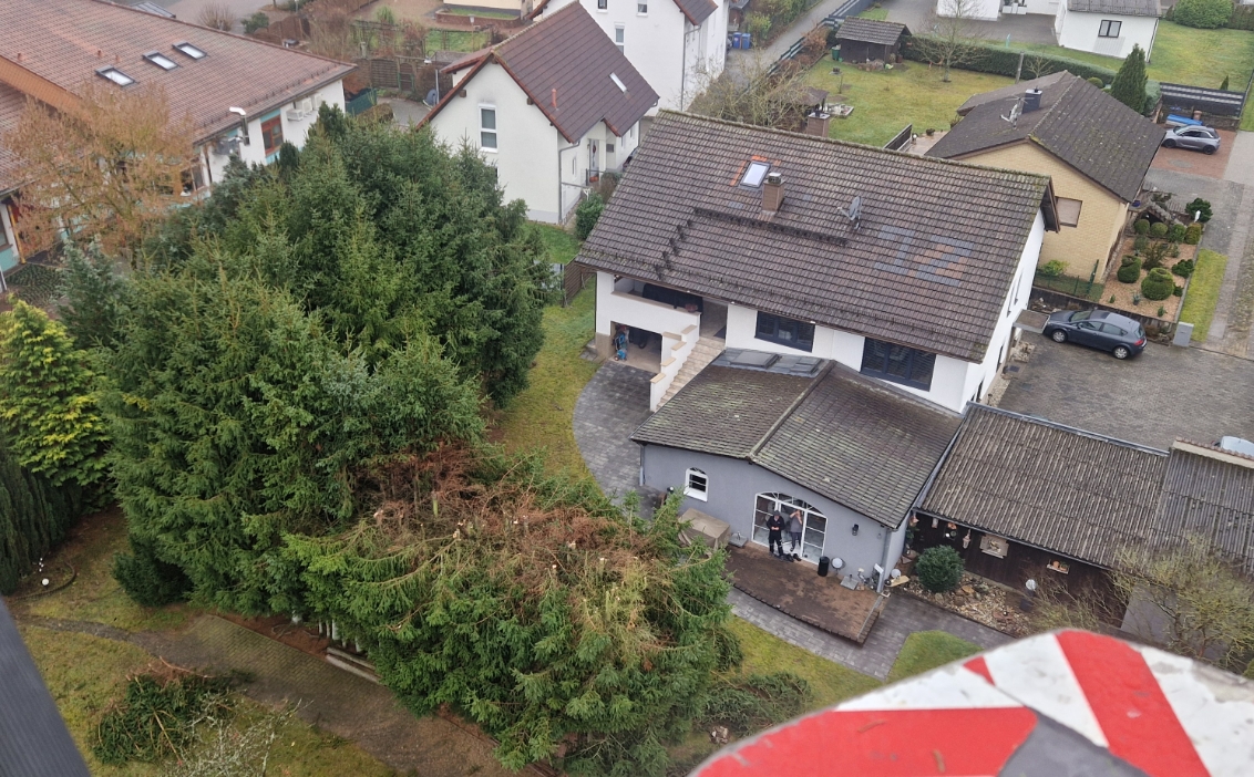 Vogelperspektive auf ein weißes Wohnhaus mit grauem Anbau, mehreren parkenden Autos und dichten Nadelbäumen im Garten.