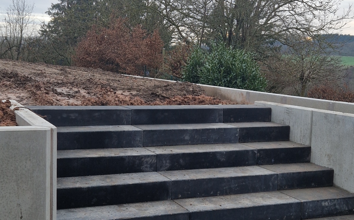 Freiliegende schwarze und graue Betontreppe mit beigen Betonstützwänden, im Hintergrund Vegetation und schlammiger Boden.