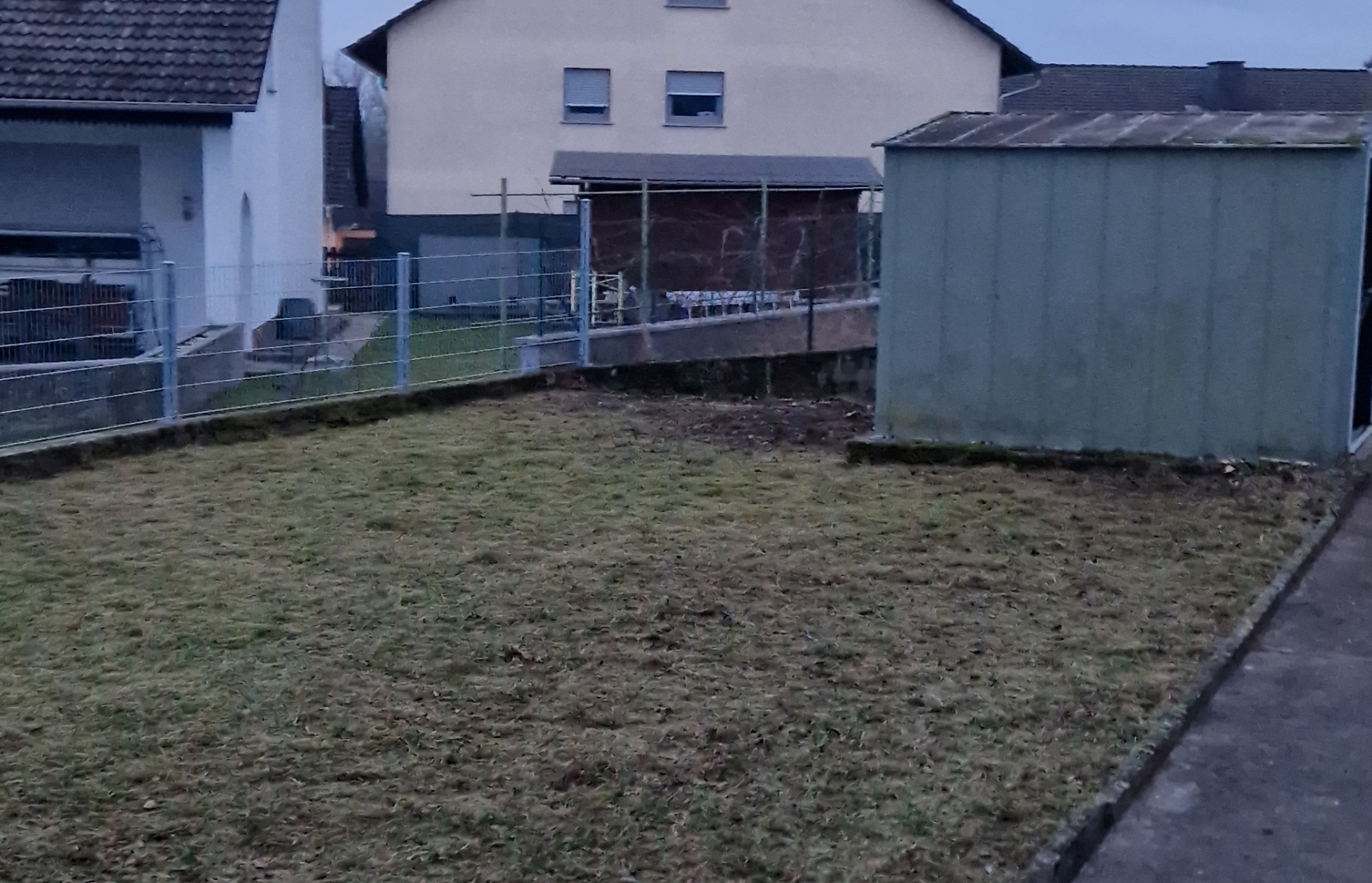 Ein eingezäunter, ungepflegter Rasen mit einem kleinen Schuppen und Häusern im Hintergrund bei trübem Wetter.