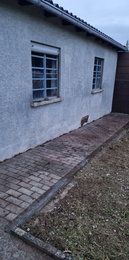 Gepflasterter Gehweg entlang einer weißen Hauswand mit zwei vergitterten Fenstern und einem kleinen beschädigten Bereich, neben einer Rasenfläche.