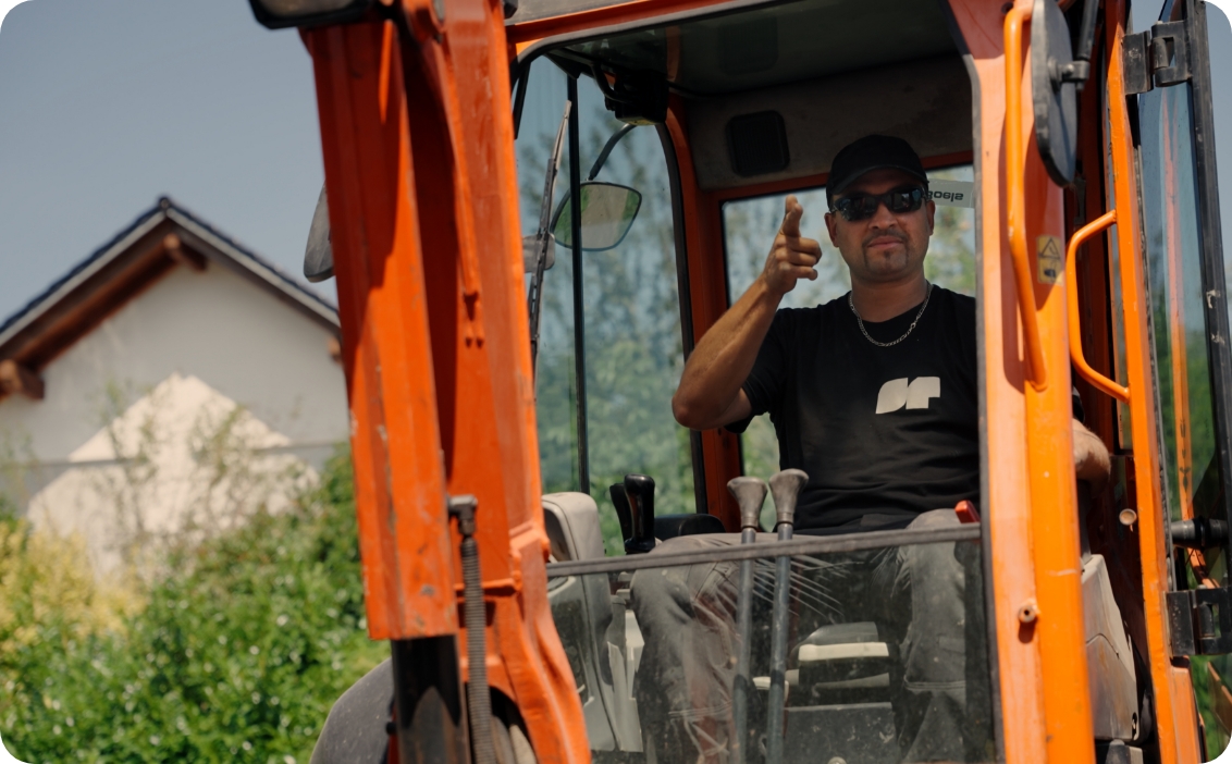Mann mit Sonnenbrille und Mütze sitzt in einem orangefarbenen Bagger und zeigt mit dem Finger zur Kamera.