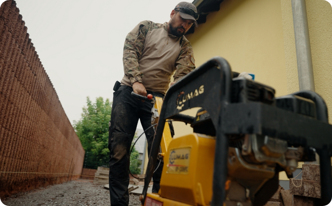 Mann in Arbeitskleidung bedient eine gelbe Bodenverdichtungsmaschine auf einer Baustelle neben einer gelben Wand.