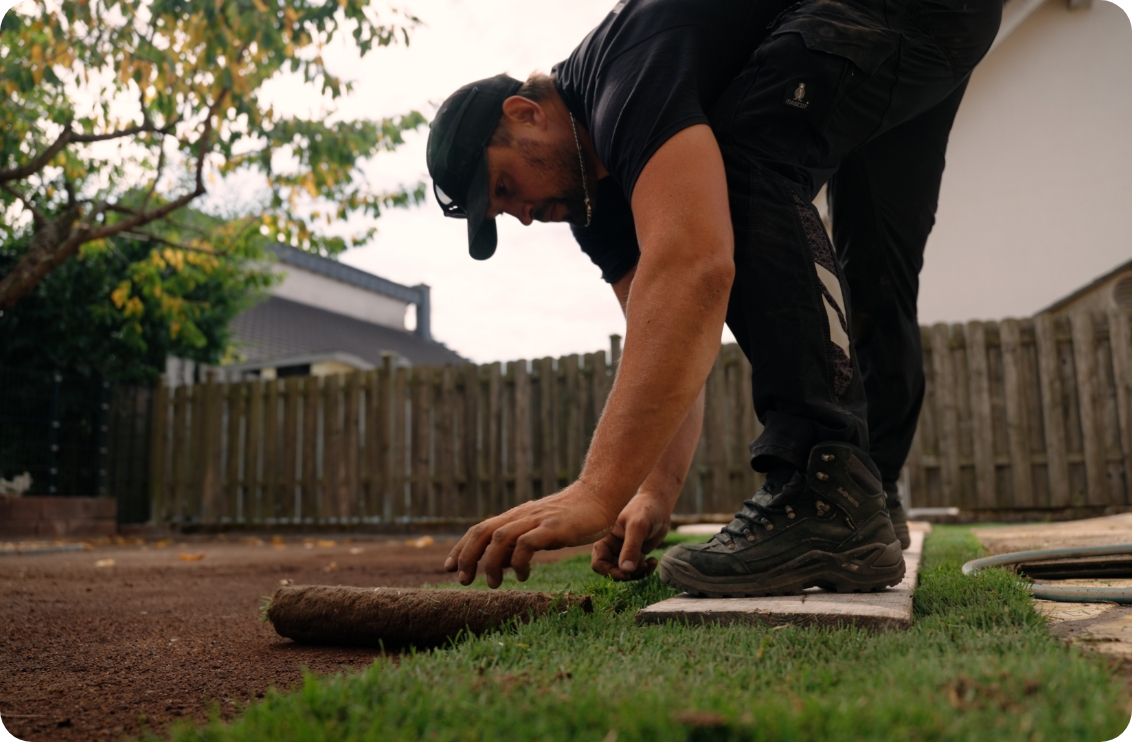 Ein Mann verlegt Rollrasen im Garten, kniet auf einer Holzplatte und legt ein Stück Gras auf den Boden.
